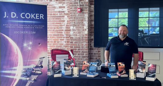 J.D. Coker at his author booth beside a banner reading Epic Space Opera in a Universe Reborn from Earth's Ashes, with Shaper Saga novels displayed